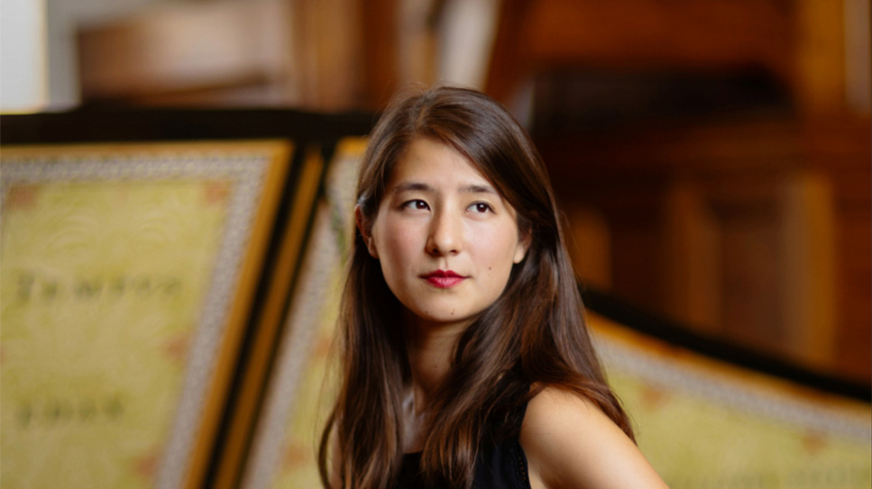 A women with long hair, sitting next to a harpsichord, looking away from the camera with a small smile.